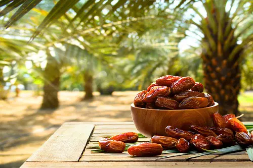 Bowl of fresh Iranian dates on a wooden table with palm trees in the background – NOVEXPORT premium dates